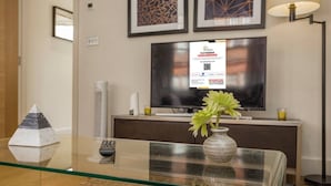 Et hotelværelse med et glasbord, en fjernbetjening, en vase med blomster, en pyramidepynt, et fladskærms-tv og indrammede billeder på væggen.