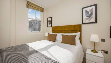A hotel room with a bed, a nightstand, a lamp, a window with a view of buildings, and two framed pictures on the wall.