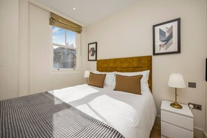 A hotel room with a bed, a nightstand, a lamp, a window with a view of buildings, and two framed pictures on the wall. - Fitzrovia 1BR Retreat | Near Soho & Tube-10 (Greater London)