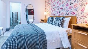 Una habitación de hotel con cama, mesita de noche, espejo, papel tapiz floral y una silla.