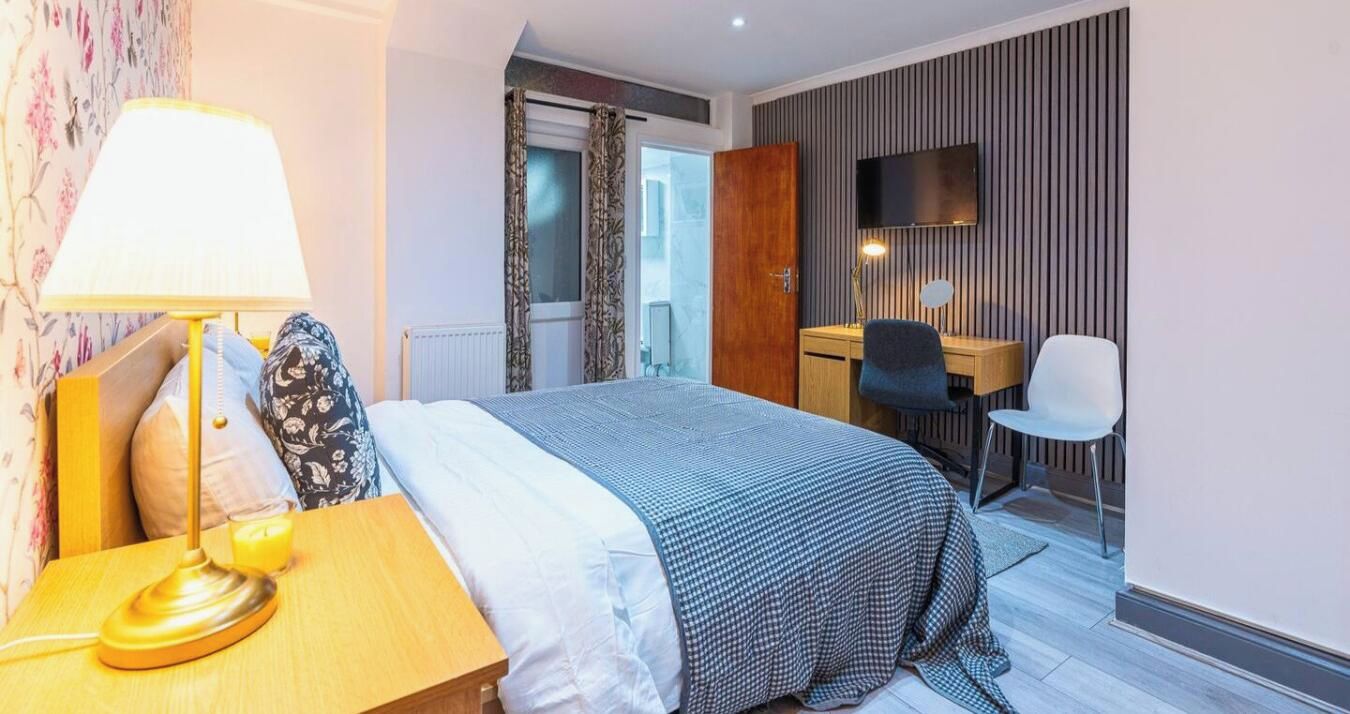 Een hotelkamer met een bed, nachtkastje, lamp, stoel, bureau en een aan de muur bevestigde televisie.