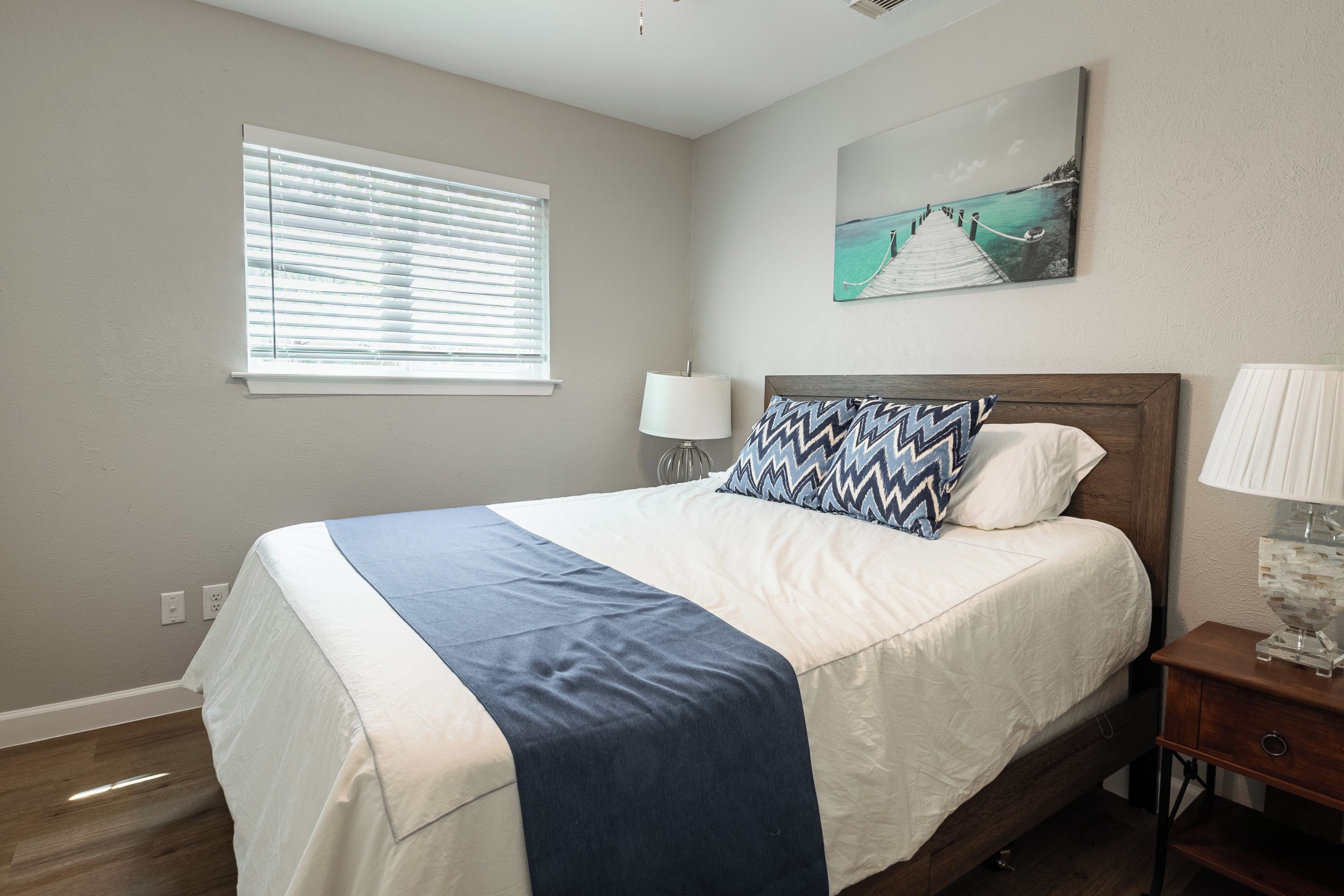 A bedroom with a bed, a nightstand, a lamp, a window with blinds, and a painting on the wall.