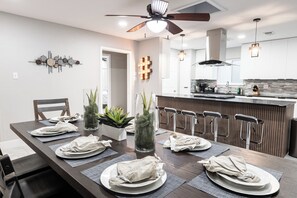 A modern dining area with a long table set for six, a kitchen in the background, and a ceiling fan. - Luxury SoCo Oasis, Private Pool, Large Covered Patio & Shuffleboard (Austin)