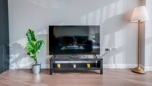 Een flatscreen televisie op een houten standaard, samen met een afstandsbediening, een kamerplant en een vloerlamp in een kamer met houten vloeren.