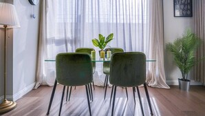 Een eetgedeelte met een glazen tafel, vier stoelen, een potplant en een wandklok.