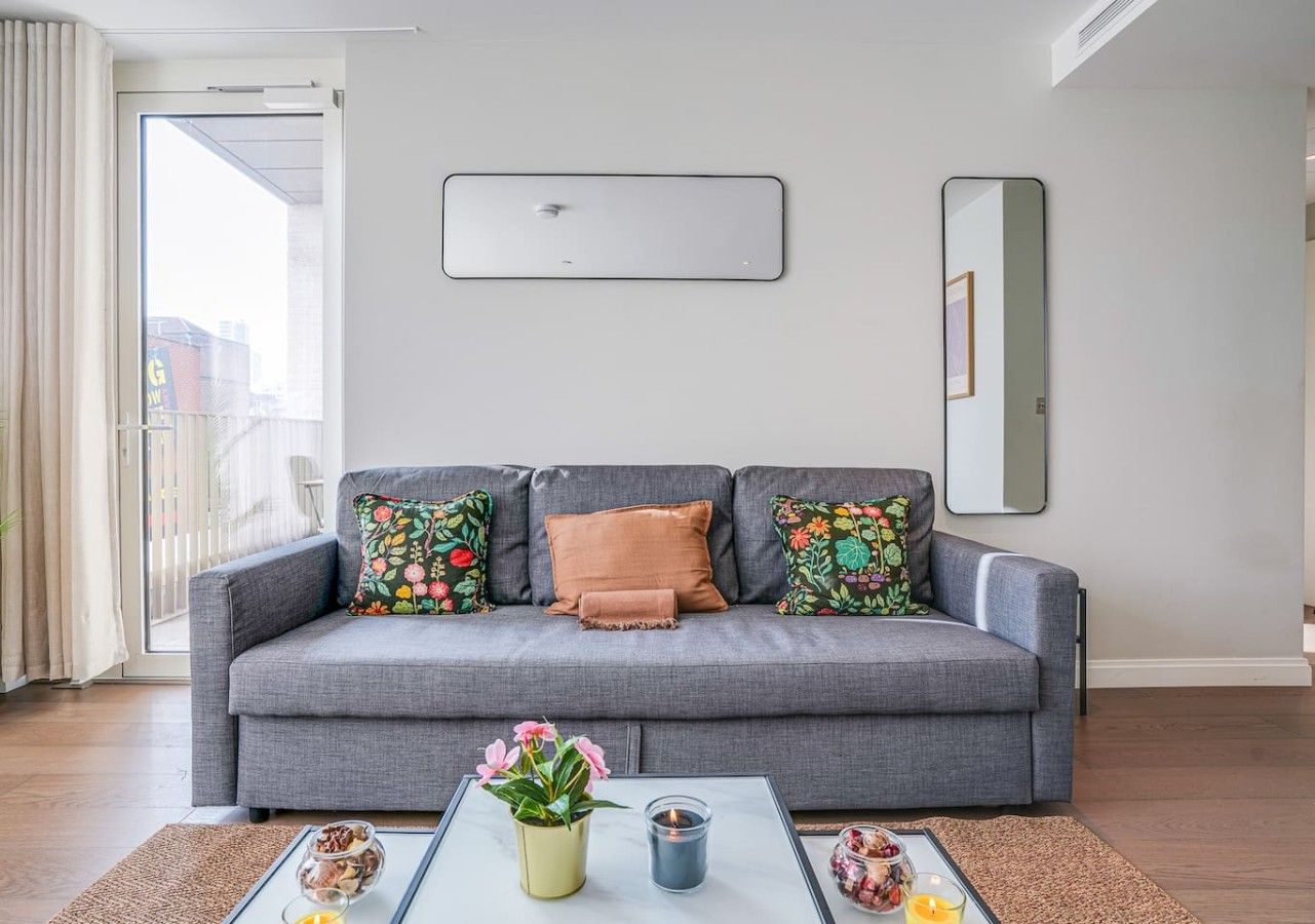 A living room with a grey sofa, colorful floral cushions, a glass coffee table with a vase of flowers, and a mirror on the wall.