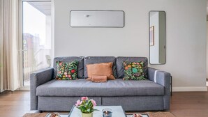 Een woonkamer met een grijze bank, kleurrijke bloemenkussens, een glazen salontafel met een bloevas en een spiegel aan de muur.