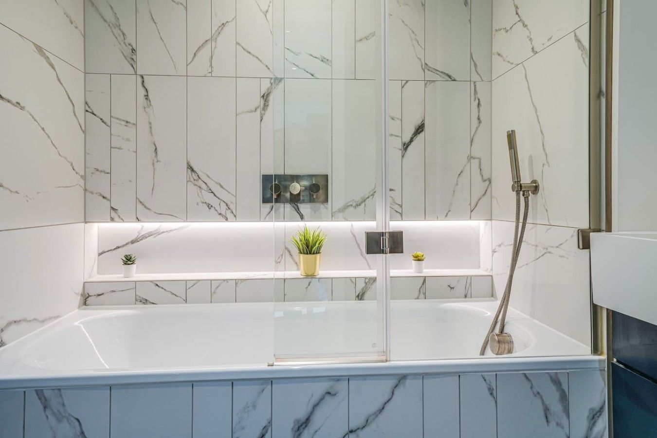 A modern bathroom with a glass-enclosed bathtub, marble walls, and a shelf with plants.