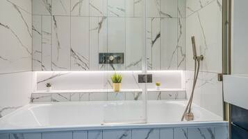 A modern bathroom with a glass-enclosed bathtub, marble walls, and a shelf with plants.