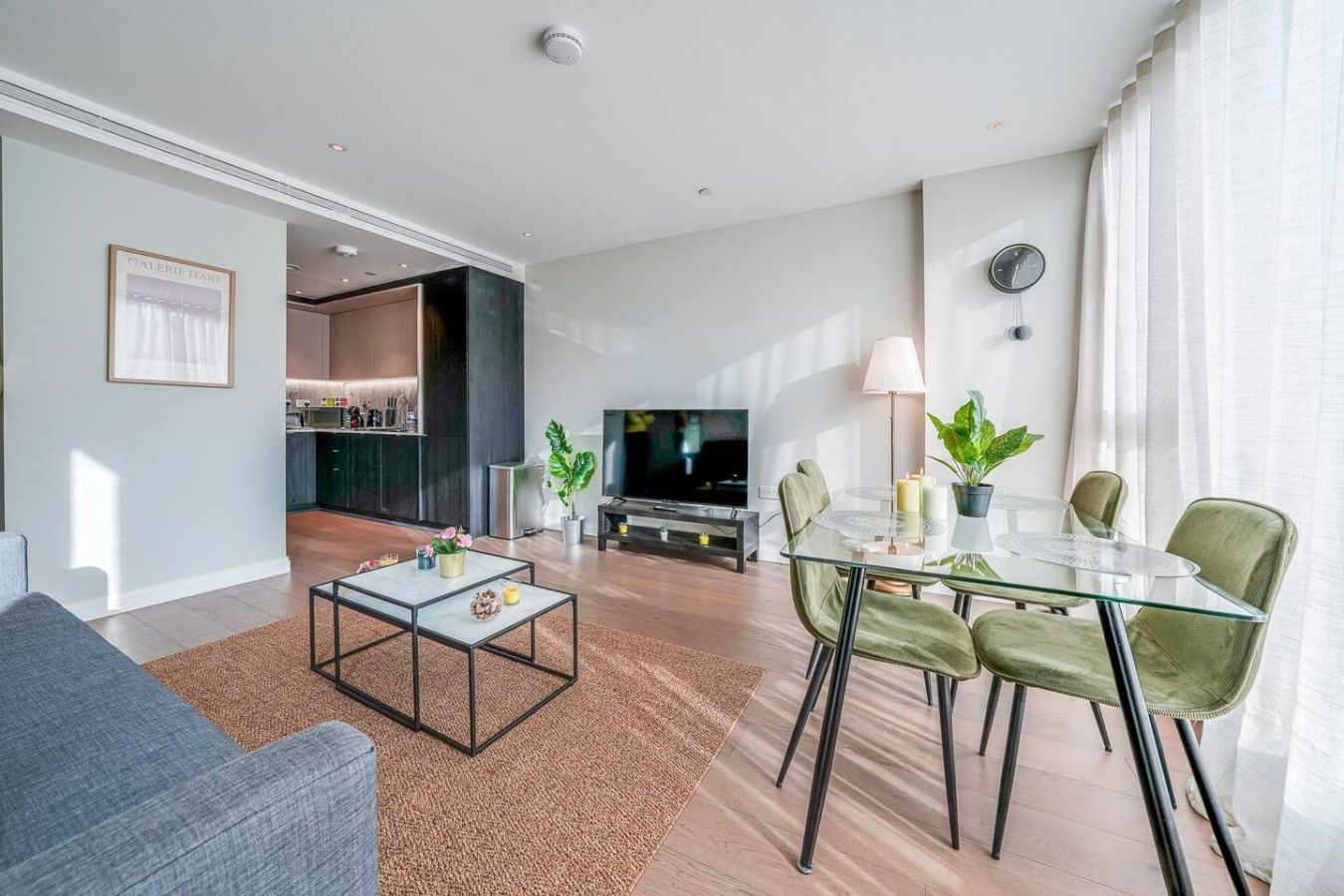 A modern living room with a glass coffee table, a sofa, a dining area with a glass table and chairs, a kitchen in the background, and a flat-screen TV.