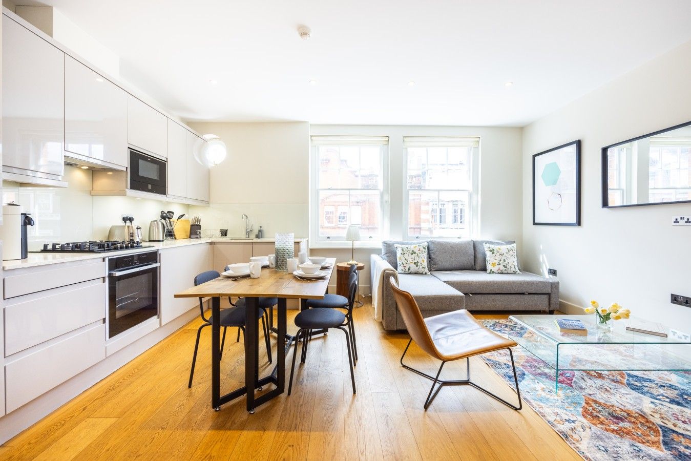 A modern kitchen with white cabinets, a dining table, and a sofa in a bright living area with large windows.