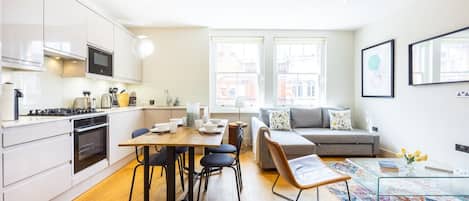 A modern kitchen with white cabinets, a dining table, and a sofa in a bright living area with large windows.