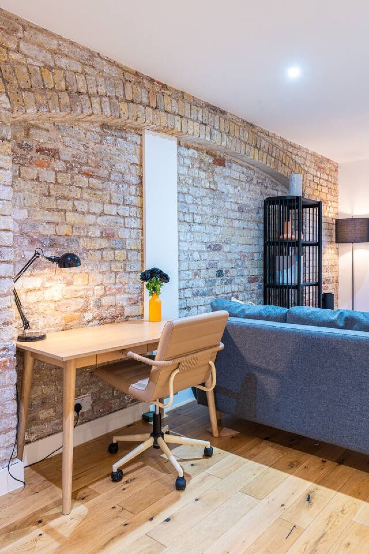 A room with a brick wall, a wooden desk with a chair, a bookshelf, and a sofa.