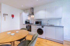 A compact kitchen with white cabinets, a built-in oven, a microwave, a washing machine, and a dining area with a round table and chairs. - Farringdon Entire Building | 4BR | Sleeps 18 (Greater London)