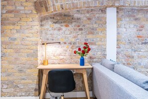 A room with a brick wall, a wooden desk with a lamp, a vase with flowers, and a grey sofa.