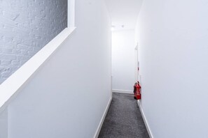 A narrow hallway with white walls, a grey carpet, and a fire extinguisher mounted on the wall.