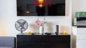 Ein modernes Wohnzimmer mit einem Flachbildfernseher an der Wand, einer schwarzen Unterhaltungseinheit darunter, einem Standventilator, einer Vase mit Blumen und einer Mikrowelle.