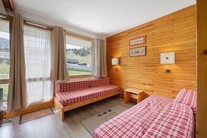 A room with a wooden wall, a bed, a bench, and a window with curtains.