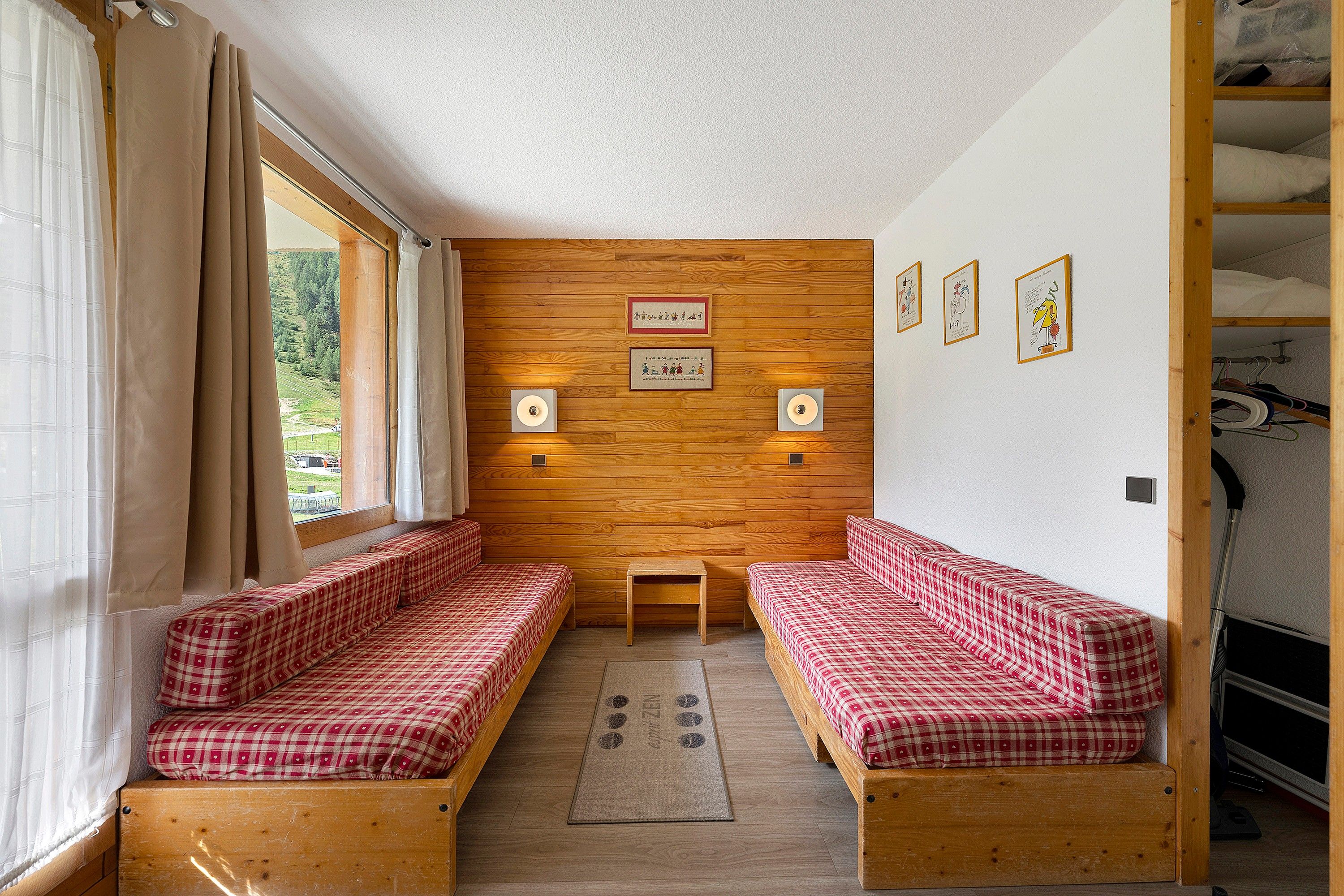 A room with wooden walls, two benches with checkered bedding, a small table, and framed pictures on the wall.