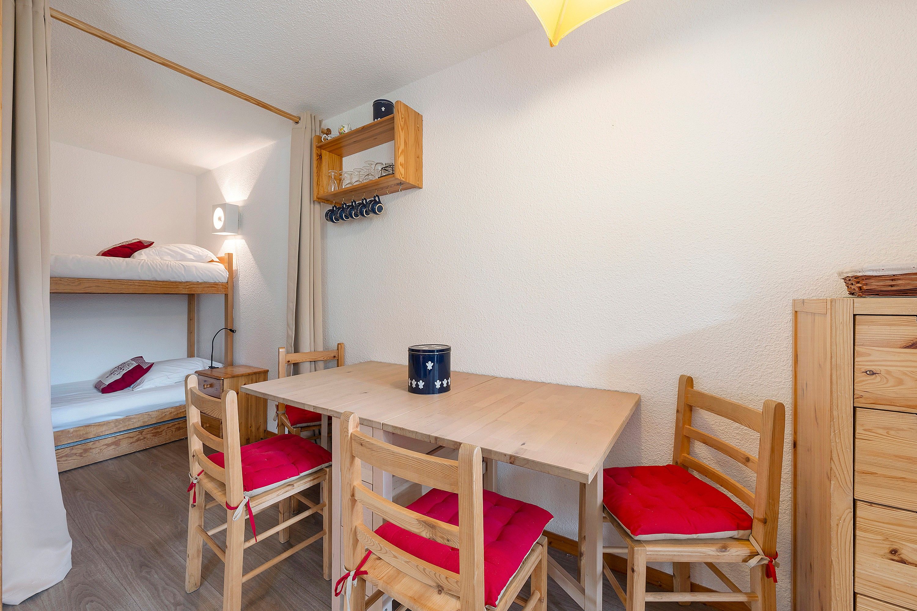 Ett rum med en våningssäng, ett träbord med stolar, en lampa och en hylla med glas.