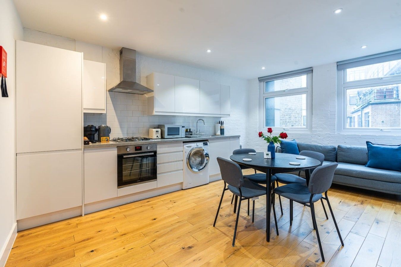 A modern kitchen with white cabinets, a built-in oven, and a washing machine. There is a dining area with a round table and chairs, and a sofa with blue cushions.