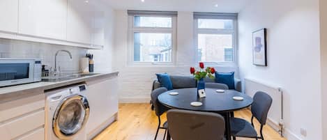 A compact kitchen with a microwave, washing machine, and a dining area with a round table and chairs.