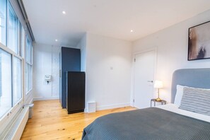 A bedroom with a bed, a nightstand, a lamp, a window, and a door. - Farringdon 1BR | Sleeps 4 | Elizabeth Line| Zone 1 (Greater London)