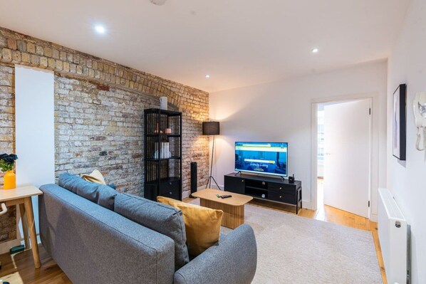 Una sala de estar con pared de ladrillo, un sofá gris, un mueble de entretenimiento negro, un televisor de pantalla plana y una mesa de centro de madera.
