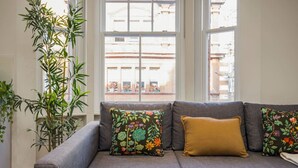 A grey sofa with floral and mustard-colored cushions, positioned in front of a window with a view of buildings. - Fitzrovia 1BR Retreat | Near Soho & Tube-8 (Greater London)