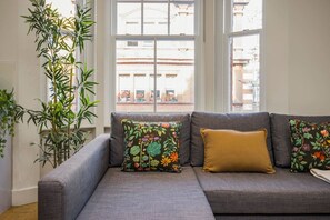 Een grijze bank met kussens in bloemen- en mosterdkleur, geplaatst voor een raam met uitzicht op gebouwen.