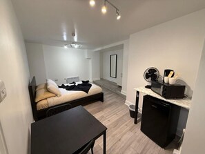 A compact room with a bed, a small table, a kitchenette with a microwave, and a fan.