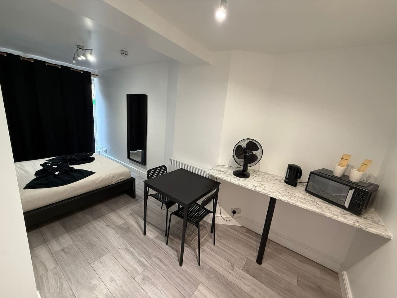 Habitación pequeña con cama, una mesita con sillas, microondas, ventilador y cafetera.