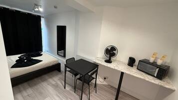 Habitación pequeña con cama, una mesita con sillas, microondas, ventilador y cafetera.