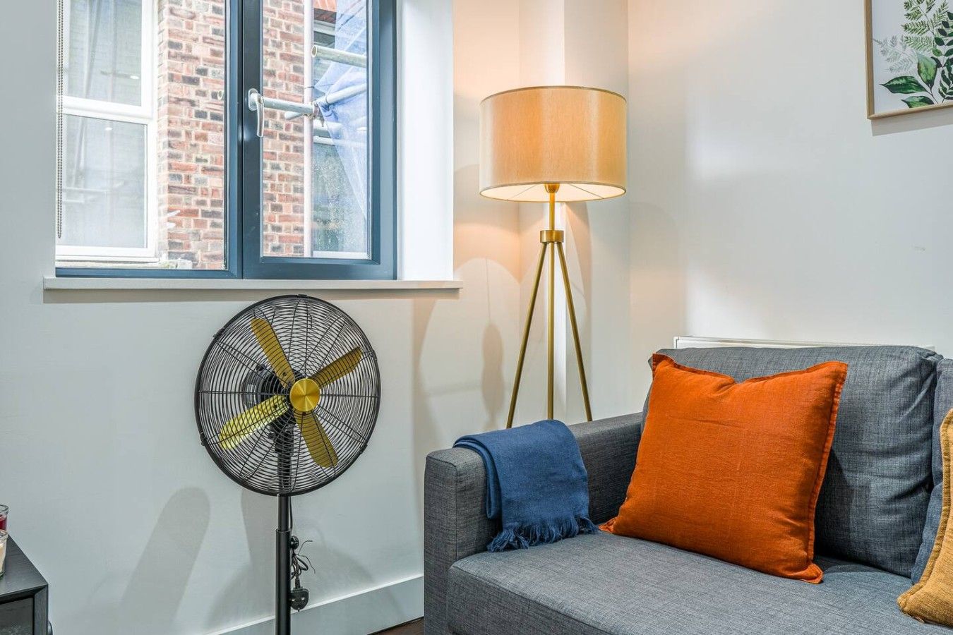 Ein Zimmer mit einem grauen Sofa, einem orangefarbenen Kissen, einem Standventilator und einer Stehlampe.