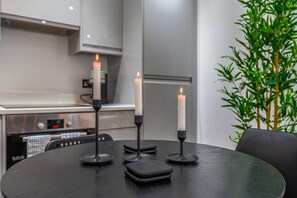 A round table with three candles in holders, a kitchen area with cabinets and appliances, and a green plant. - Earl`s Court Gem | Sleeps 6 | Lift & Tube (Greater London)