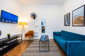 Ein modernes Wohnzimmer mit Flachbildfernseher, Standventilator, blauem Sofa, kleinem Tisch und Stuhl.