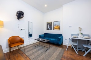 A modern living room with a blue sofa, a brown leather chair, a coffee table, and a dining area with a table and chairs. - Earl`s Court Residence | Elegant 1BR | London (Greater London)