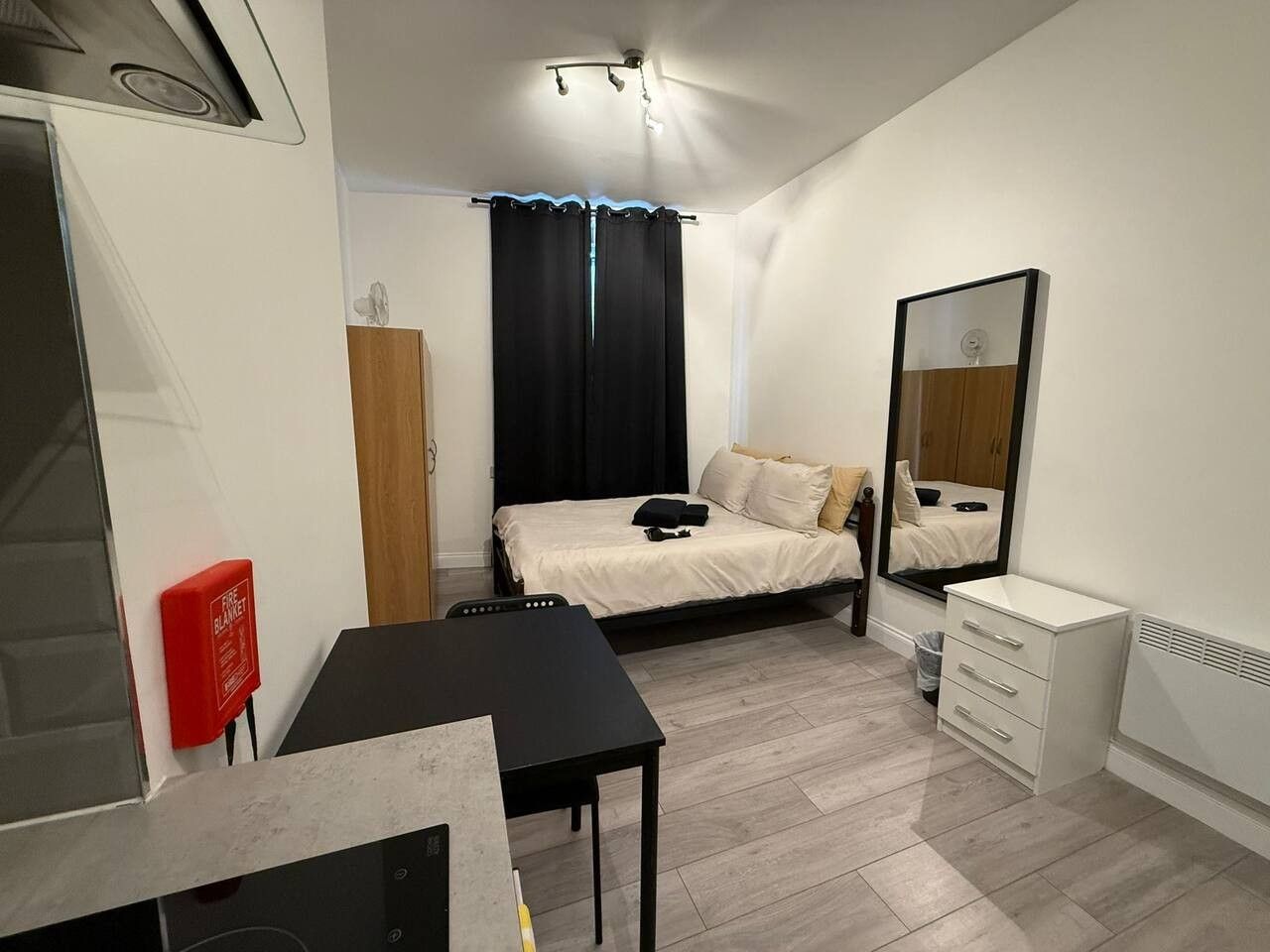 A compact bedroom with a bed, a bedside table, a mirror, a wardrobe, and a small desk area.