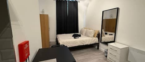 A compact bedroom with a bed, a bedside table, a mirror, a wardrobe, and a small desk area.