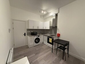 A compact kitchen with white cabinetry, a washing machine, a microwave, and a small dining area with a table and chair. - Modern Room | Minutes from Central London (Greater London)