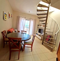 Quarto com mesa de jantar redonda, cadeiras, televisão, escada em espiral e vista para o exterior.