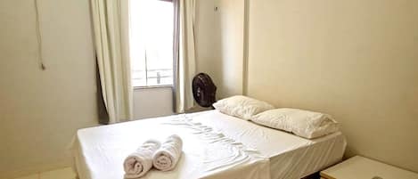 A hotel room with a bed, a bedside table, a window with curtains, and an air conditioning unit.