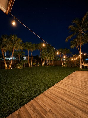 A nighttime outdoor area with a wooden deck, string lights, and palm trees.