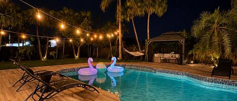 A swimming pool with lounge chairs and swan-shaped floats, surrounded by palm trees and string lights.