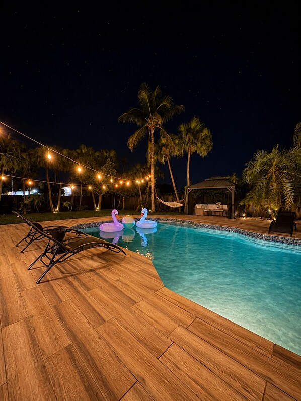 A swimming pool with lounge chairs and swan-shaped floats, surrounded by palm trees and string lights.