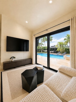 A modern living room with a flat-screen TV, a dark wooden console, a black ottoman, and a large window overlooking a swimming pool.