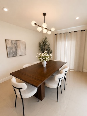 Une salle à manger avec une longue table en bois, des chaises blanches, un vase rempli de fleurs, un tableau accroché au mur et un luminaire au plafond.