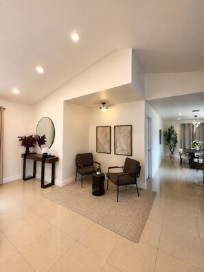 A spacious living room with a high ceiling, recessed lighting, a coffee table, two armchairs, a side table with a plant, and artwork on the wall.