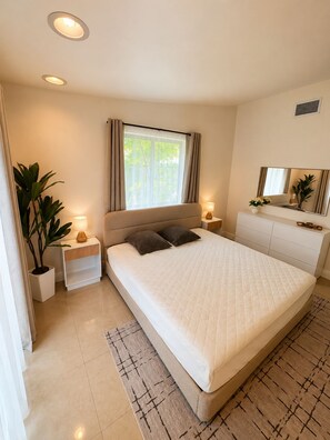 Quarto moderno com cama grande, luminárias de cabeceira, mesa de cabeceira, planta em vaso e janela com cortinas.