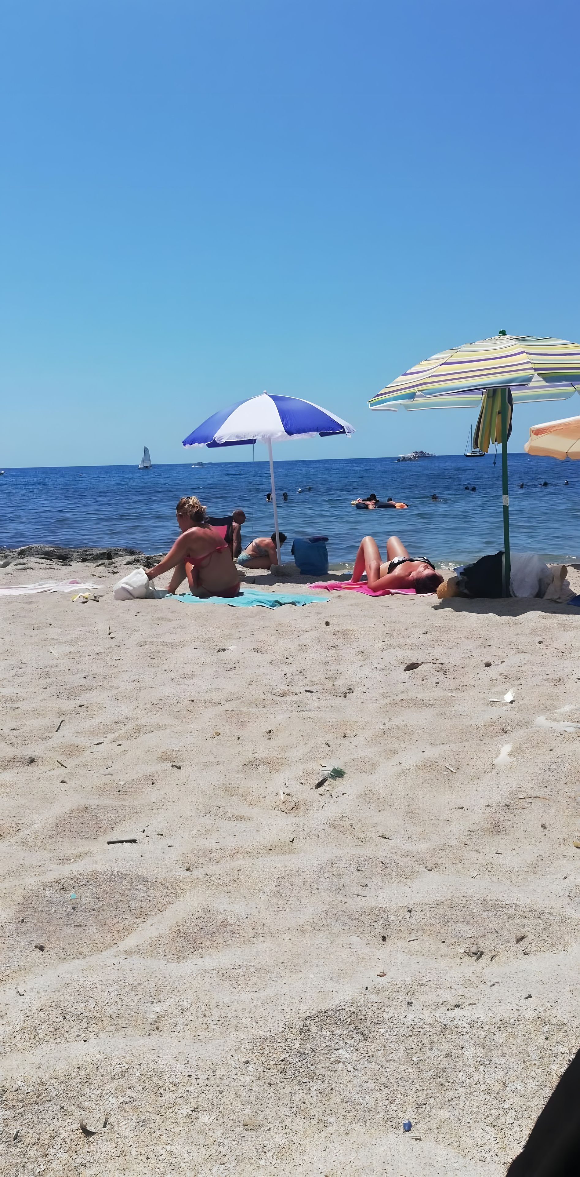 Una spiaggia con persone che prendono il sole e nuotano, una barca a vela in lontananza e un cielo azzurro limpido.